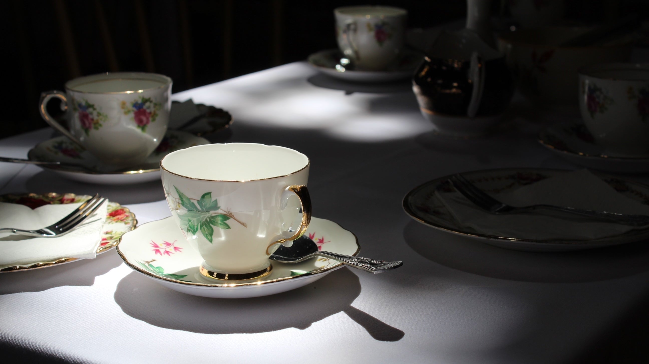 A vintage china teacup and saucer set with a delicate green leaf pattern and gold trim, sits on a white tablecloth, bathed in a single spotlight. A silver spoon rests in the saucer. In the background, other out-of-focus teacups and plates are visible.