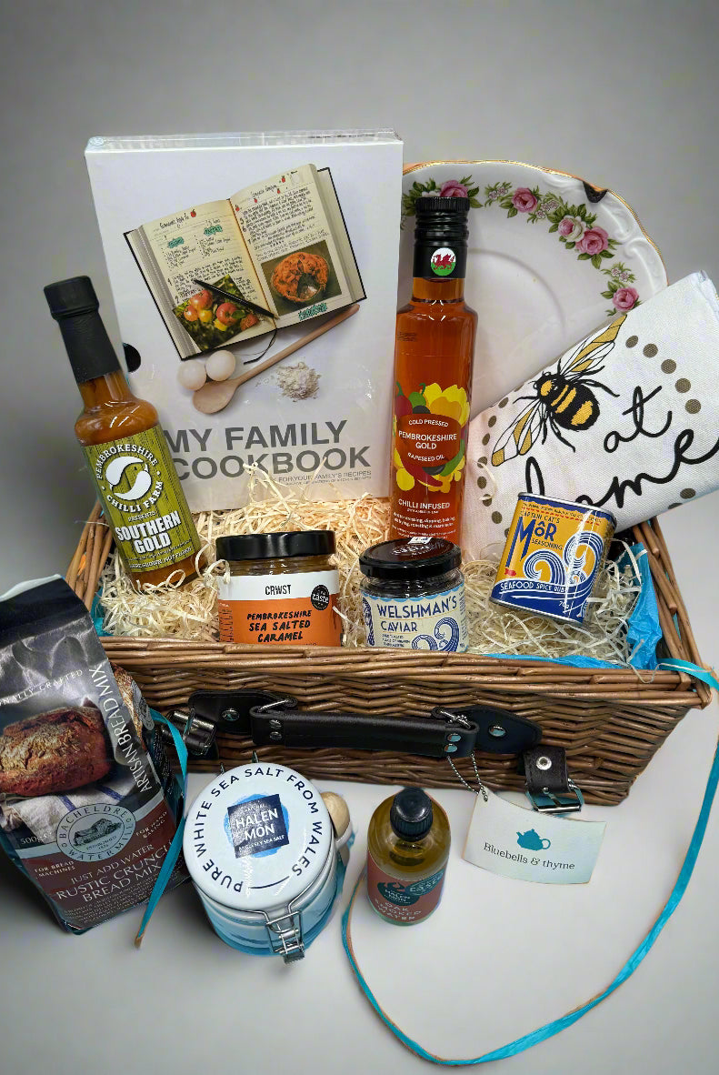 Bluebells and Thyme wicker hamper basket filled with an assortment of gourmet food items, including salt, oil, caramel, caviar, bread mix, a book, and a vintage cake plate.