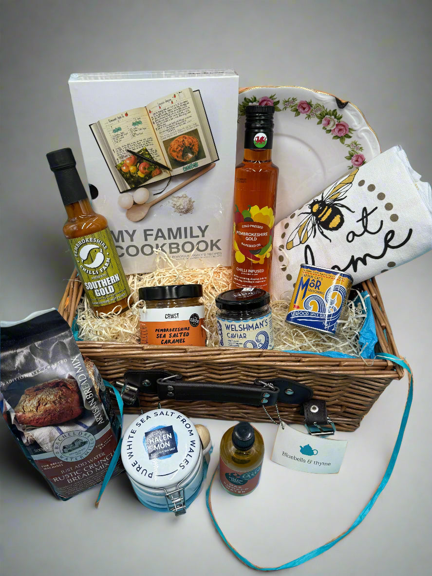 Bluebells and Thyme wicker hamper basket filled with an assortment of gourmet food items, including salt, oil, caramel, caviar, bread mix, a book, and a vintage cake plate.