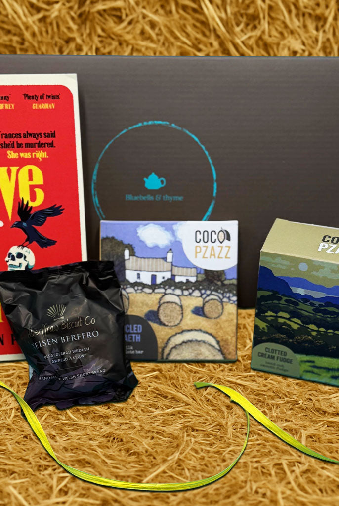 Bluebells and Thyme Just Because gift box containing a book, a handcrafted bookmark, a candle, and a box of shortbread cookies, all displayed on a straw surface.