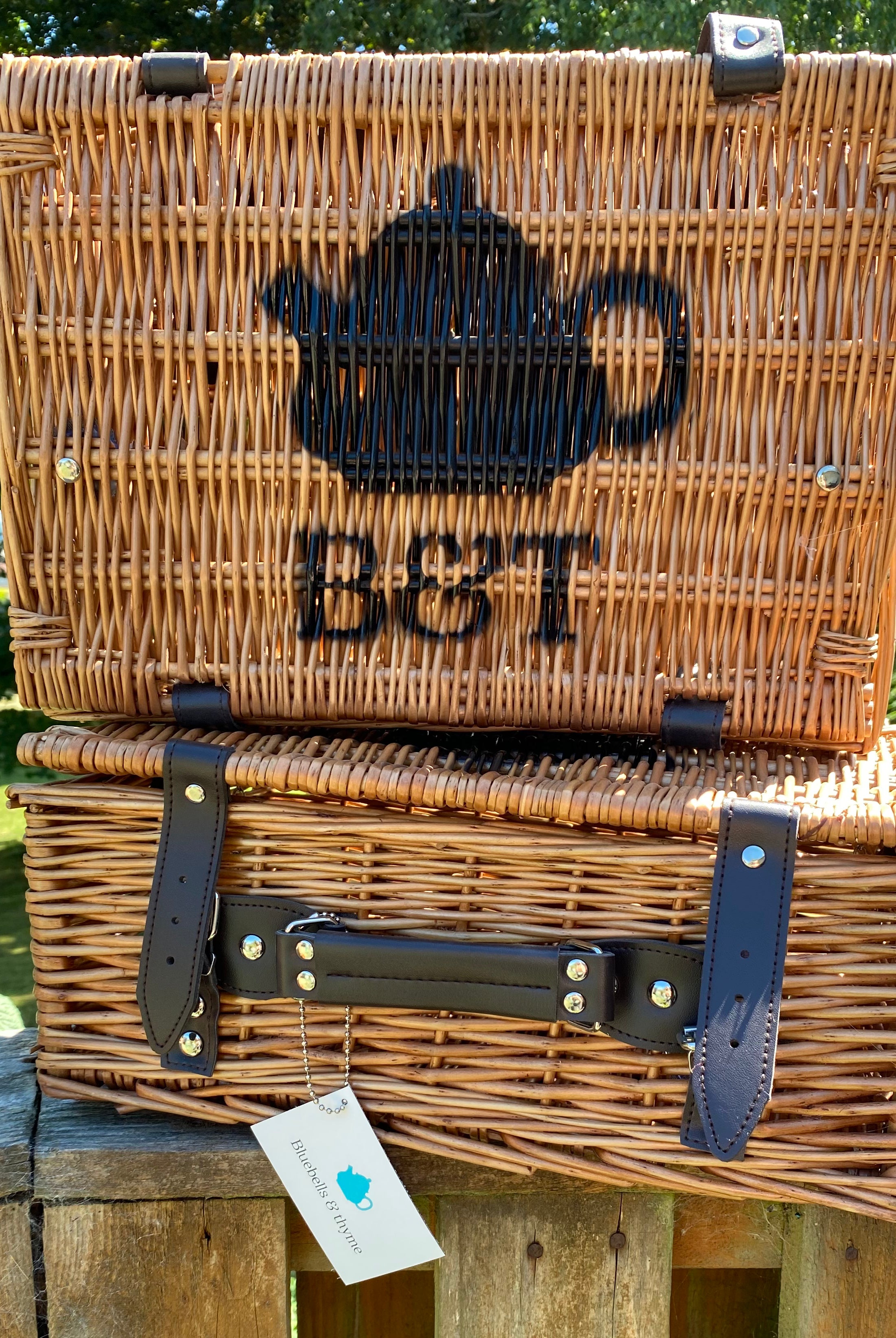 Bluebells & Thyme branded wicker hampers, stacked outdoors in sunlight. Features the distinctive stencilled teapot logo.