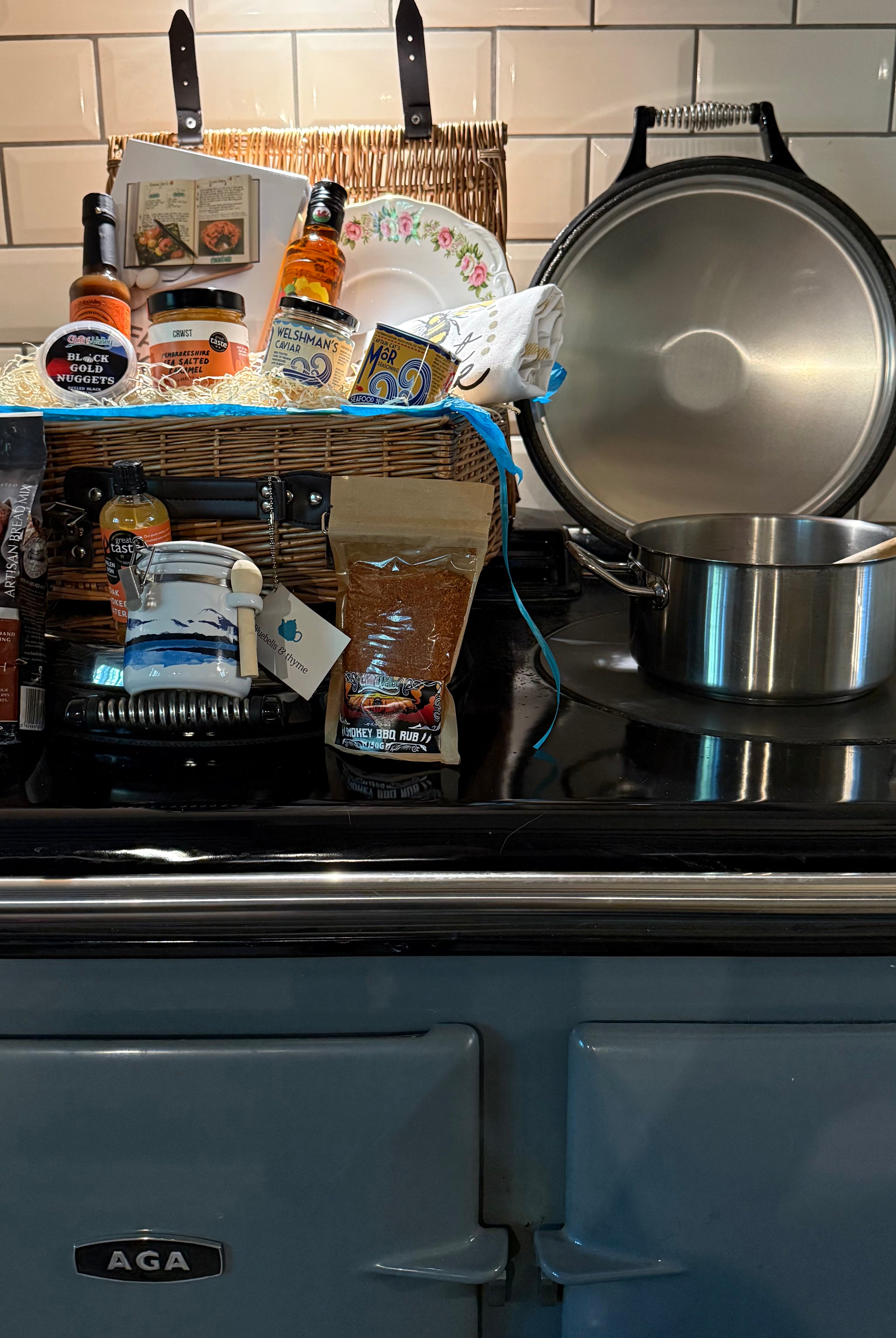 Bluebells and Thyme wicker hamper basket filled with an assortment of gourmet food items, including salt, oil, caramel, caviar, bread mix, a book, and a vintage cake plate.