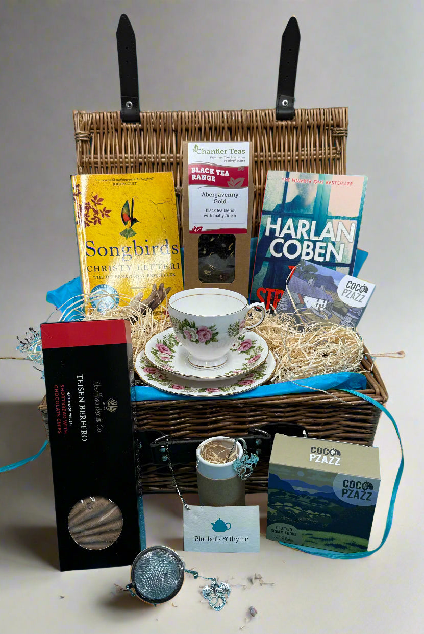 Bluebellls and thyme gift hamper set containing a vintage china cup and side plate, an assortment of books, and various tea-related items, all displayed in an open wicker basket.