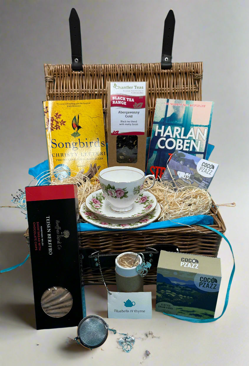 Bluebellls and thyme gift hamper set containing a vintage china cup and side plate, an assortment of books, and various tea-related items, all displayed in an open wicker basket.
