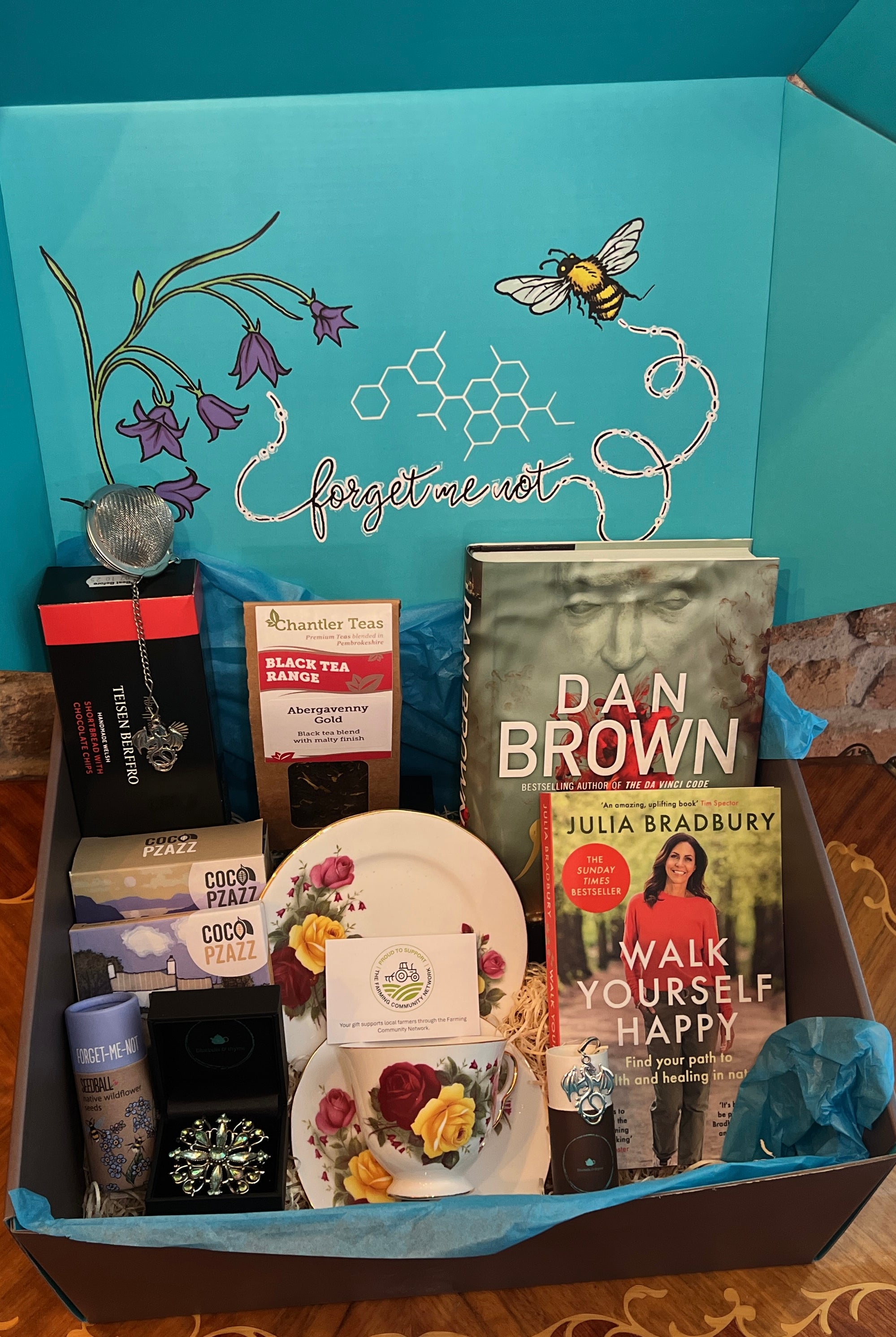 Gift hamper from bluebells and thyme containing various items including books, artisan welsh food and a vintage necklace, with a decorative blue lid featuring floral and bee designs.