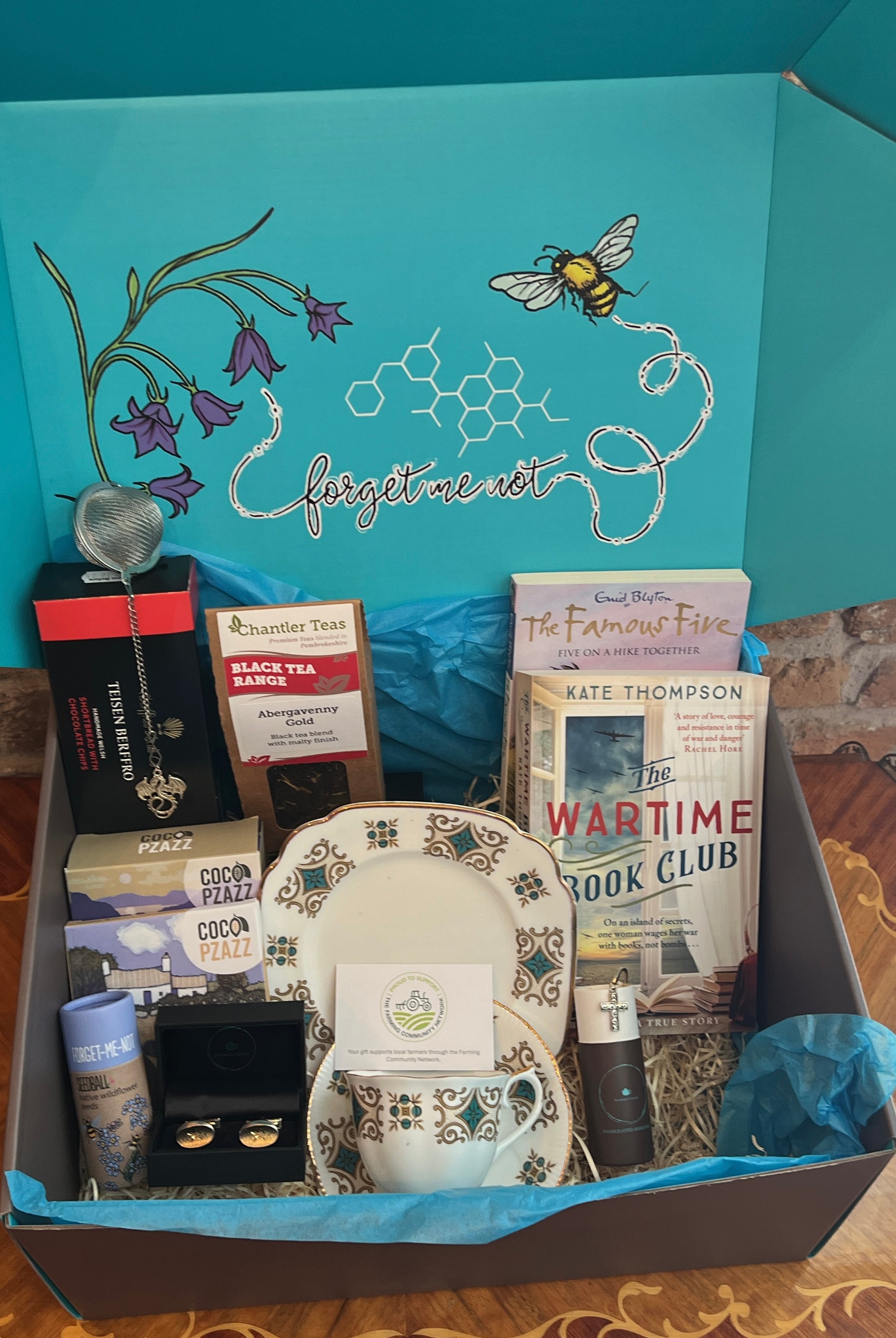 Gift Hamper by bluebells and thyme with various items including books, artisan food and vintage jewelry on a wooden surface.