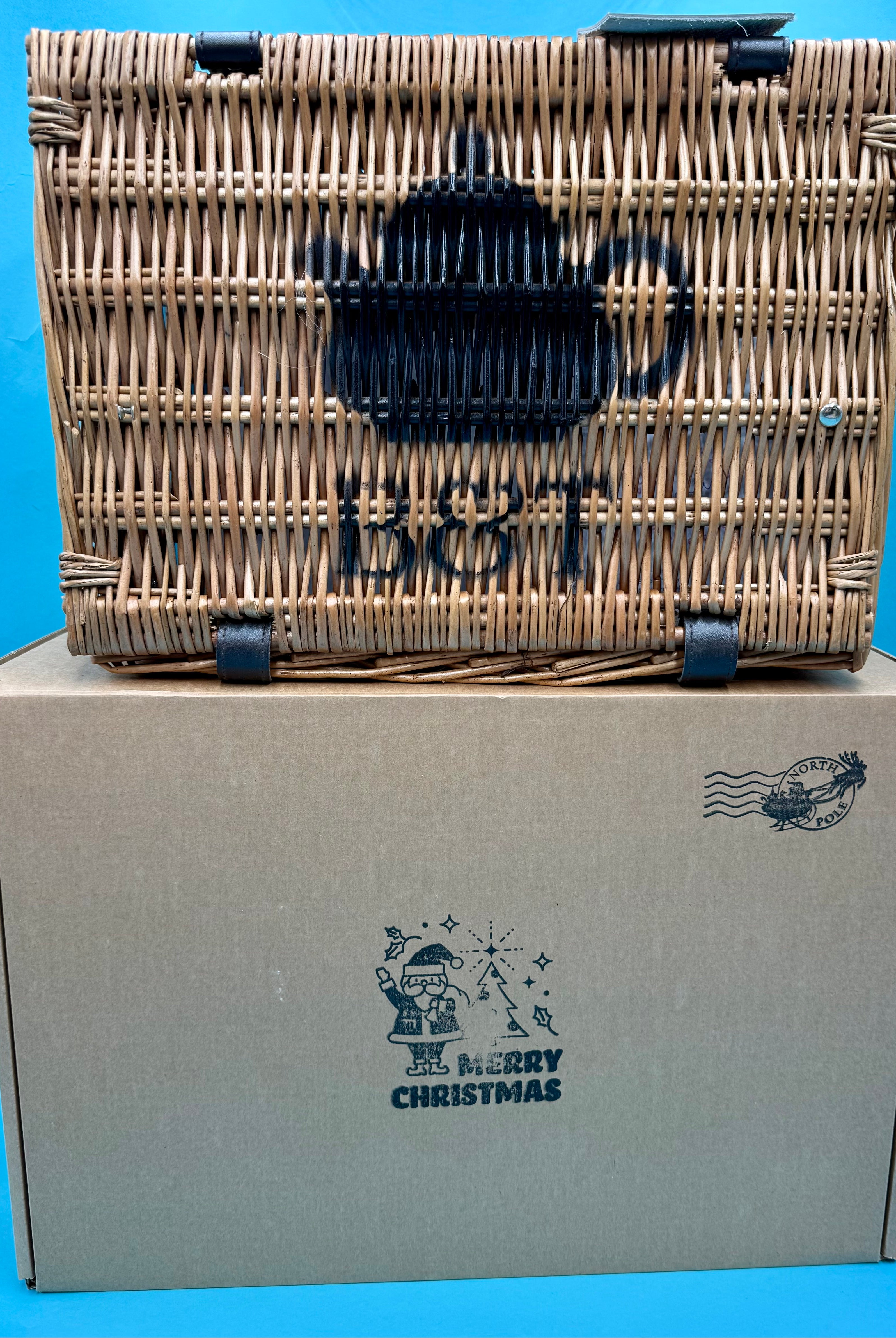 Bluebells and Thyme Wicker Hamper on top of a cardboard box with blue Christmas graphics on a blue background