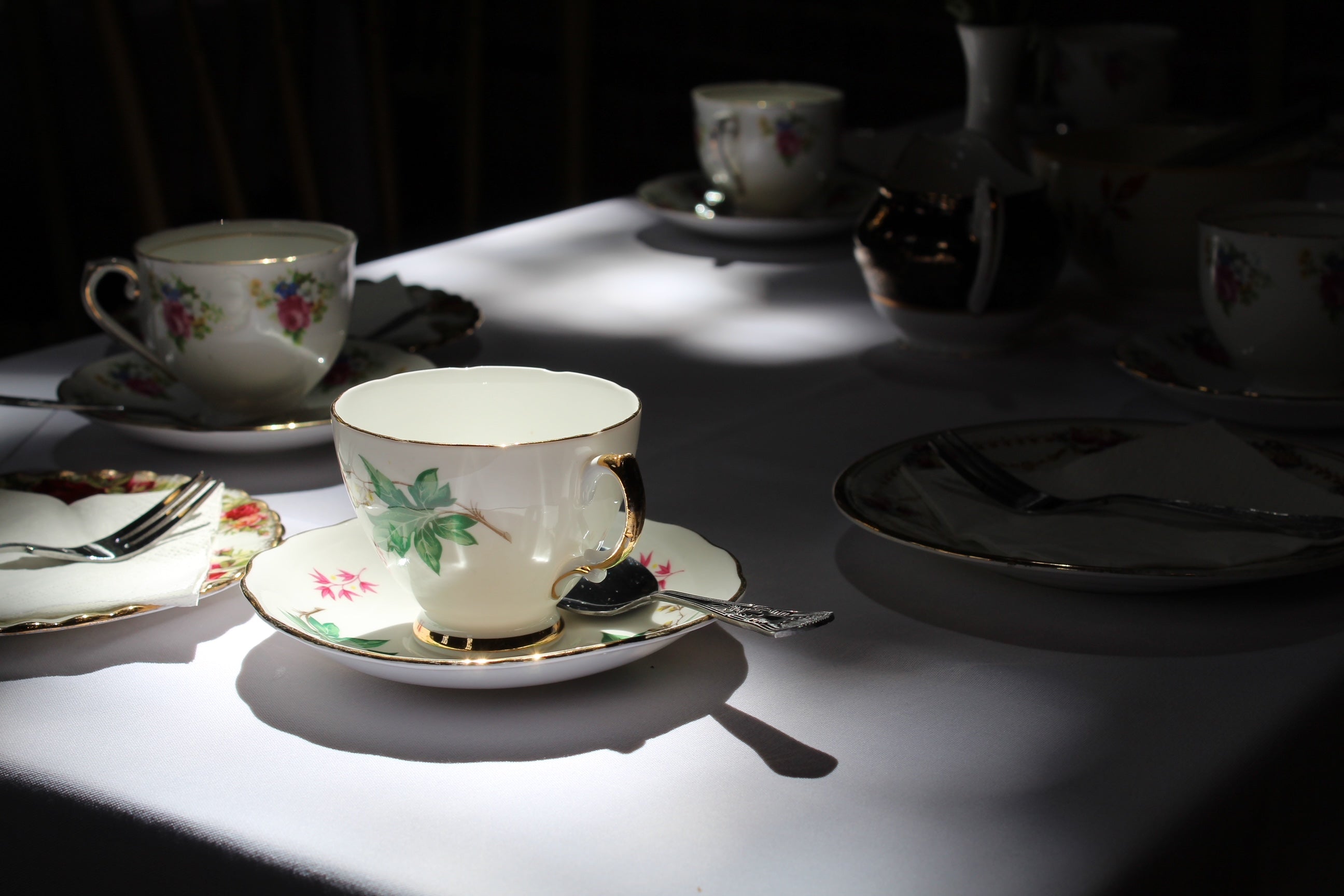 A vintage china teacup and saucer set with a delicate green leaf pattern and gold trim, sits on a white tablecloth, bathed in a single spotlight. A silver spoon rests in the saucer. In the background, other out-of-focus teacups and plates are visible.