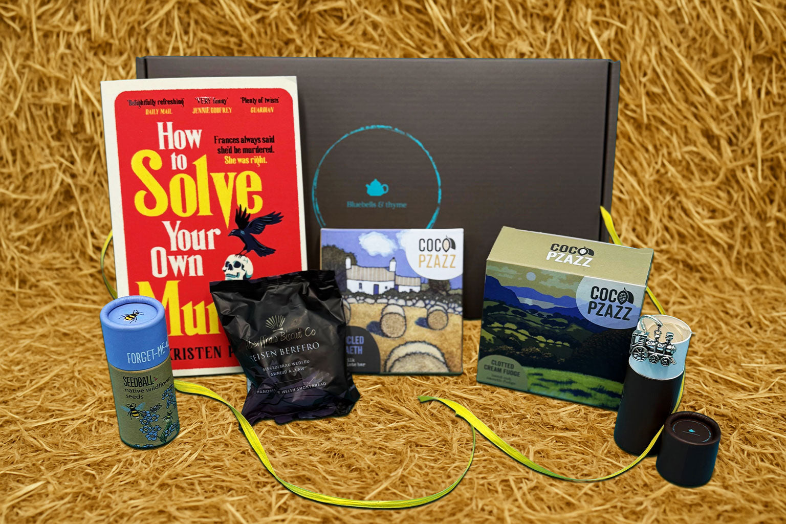 Bluebells and Thyme Just Because gift box containing a book, a handcrafted bookmark, a candle, and a box of shortbread cookies, all displayed on a straw surface.