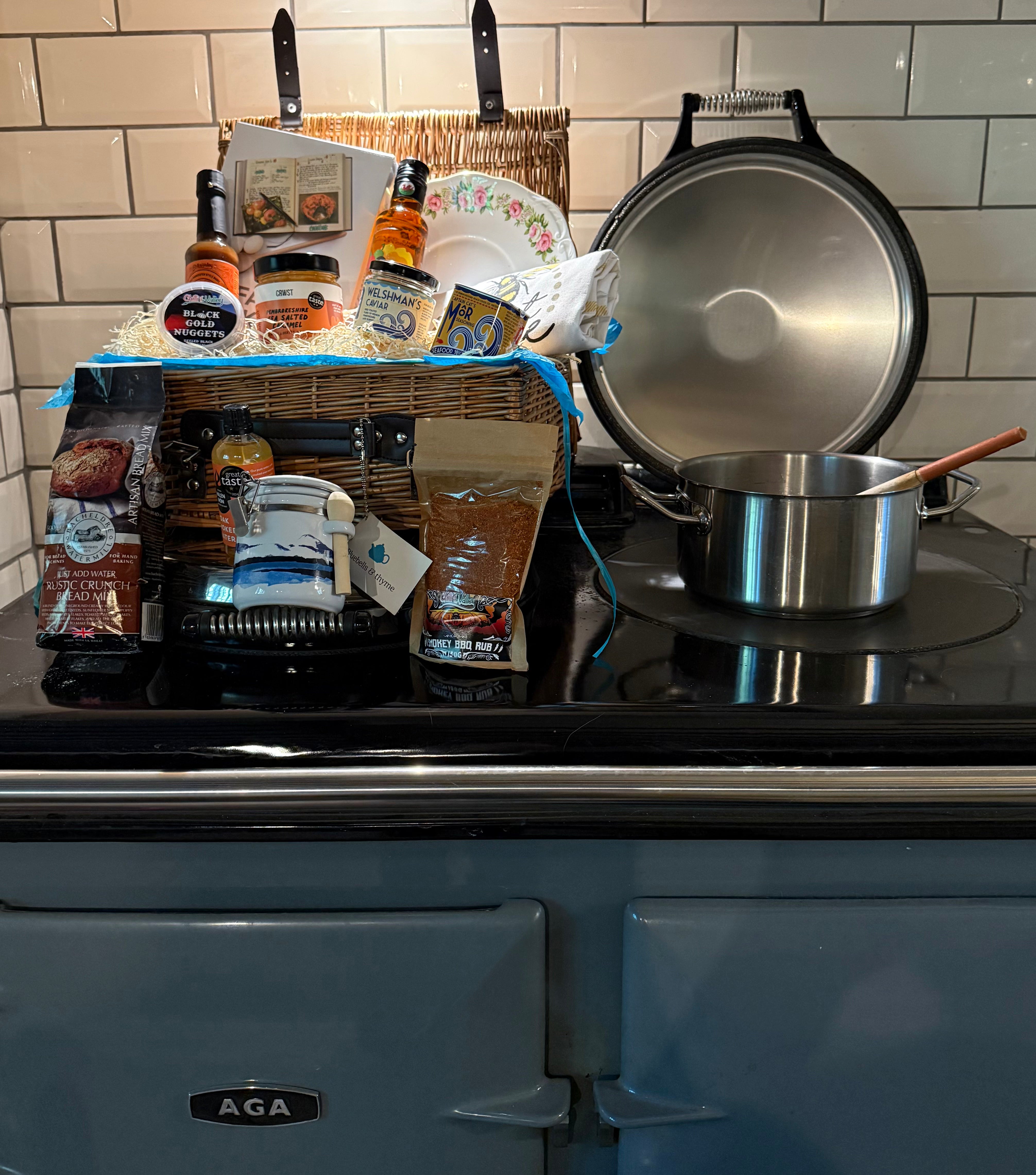 Bluebells and Thyme wicker hamper basket filled with an assortment of gourmet food items, including salt, oil, caramel, caviar, bread mix, a book, and a vintage cake plate.