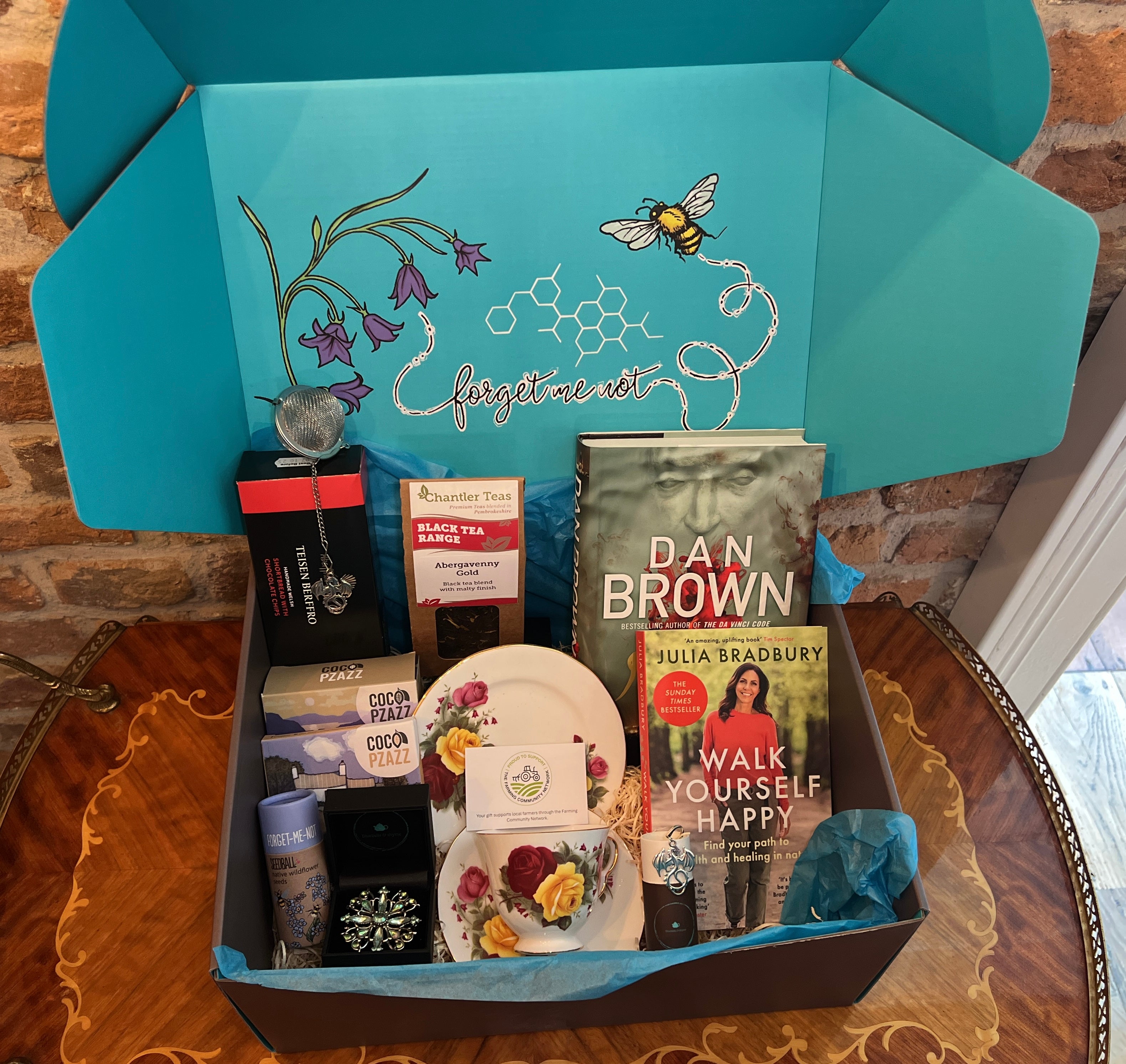 Gift hamper from bluebells and thyme  containing various items including books, artisan welsh food and a vintage necklace, with a decorative blue lid featuring floral and bee designs.