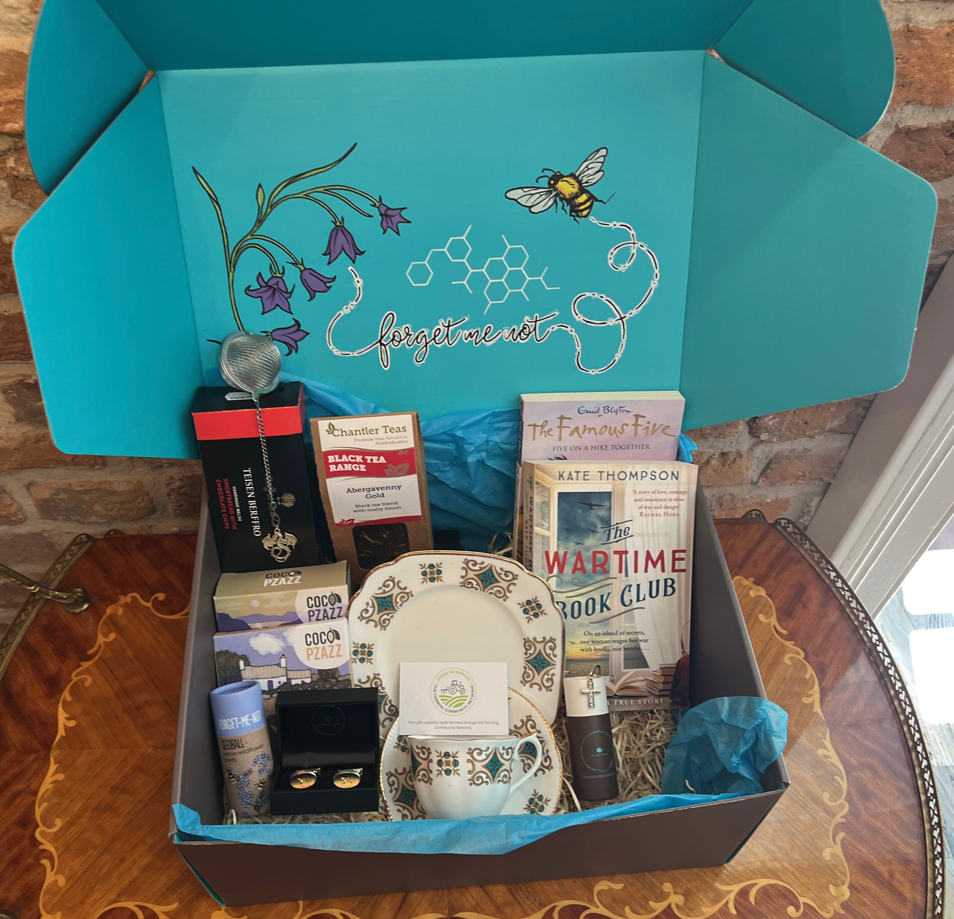 Gift Hamper by bluebells and thyme with various items including books, artisan food and vintage jewelry on a wooden surface.